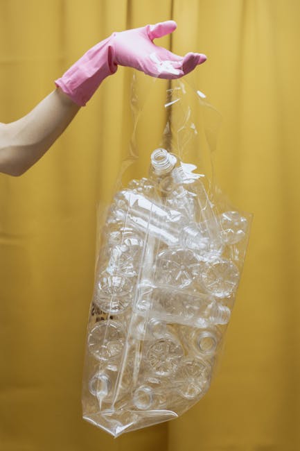 A person wearing a pink rubber glove is holding a clear plastic bag filled with used plastic bottles and containers, positioned in front of a yellow background. The bottles inside the bag are of various shapes and sizes, with some visibly crushed or deformed, and the bag appears to be partially transparent. The scene suggests the disposal or collection of plastic waste, aligned with waste removal or rubbish clearance services. The focus is on the handling of recyclable plastics, which are often part of an alternative waste management process outside regular council rubbish collection. The environment appears to be indoors, possibly in a setting related to waste sorting or recycling, emphasizing the importance of proper disposal and segregation of plastic rubbish by companies like House Clearance Bromley. The overall composition highlights the importance of responsible waste handling and the role of private collection services in managing household plastic waste.