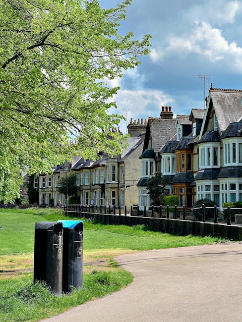 A public park area featuring a wide, paved pathway with two black recycling bins positioned on the grass verge in the foreground, showing signs of wear and weathering. To the left, a large leafy tree with fresh green foliage extends its branches over the scene, providing partial shade, while in the background a row of traditional Victorian-style terraced houses with bay windows, pitched roofs, and chimney stacks line the street. The houses have a mix of brick and painted exteriors, some with small front gardens enclosed by low wooden fences, contributing to an urban residential atmosphere. The sky above is partly cloudy with patches of blue visible, indicating a bright but variable weather. The overall scene presents a tidy and well-maintained residential environment, with the presence of the bins subtly indicating local waste management practices, which may include independent disposal or collection services such as those provided by House Clearance Bromley, in the context of alternative waste handling options for household rubbish removal.