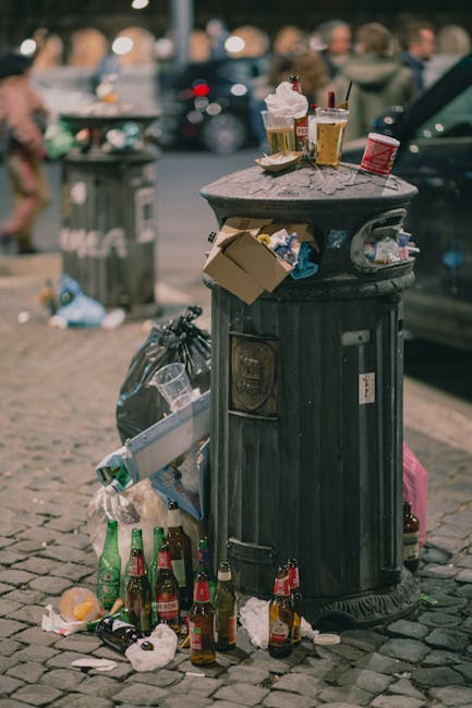 An outdoor scene showing numerous overflowing rubbish and recycling bins on a paved area near a street. The grey bin labeled for mixed paper and card is open and filled with a mixture of paper, cardboard boxes, and plastic packaging, with waste spilling onto the surrounding ground. To its right, there are black and green bins, also filled with rubbish, some contents spilling out. A large red bin is visible further right with more waste on top. Behind the bins, there is a blue metal railing, separating the waste area from a commercial building with a yellow storefront and large glass windows, which has signage and shop displays. A parked silver car is adjacent to the bins, and a tree with a bare trunk is partially visible on the left side of the image. The background features a multi-story building with blue-tinted windows and scaffolding, indicating ongoing construction or maintenance work. The scene is illuminated by natural daylight, emphasizing the cluttered and unmanaged appearance of the waste collection area, which could suggest a need for private rubbish removal services like those offered by House Clearance Bromley.