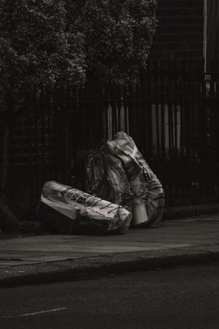 On a paved sidewalk adjacent to a black metal fence and a green leafy hedge, there are three large, tightly filled waste bags made of black plastic and white plastic material. The bags appear to contain household rubbish or garden waste, with some text or branding visible on the white bag. They are positioned close to the curb, with two standing upright and one lying against them, all slightly leaning to one side. The environment appears to be an outdoor residential area, with faint outlines of trees and a building or structure visible beyond the fence. The scene is captured in natural daylight with soft shadows, indicating an overcast or partly cloudy sky. This setting aligns with private rubbish collection or on-site clearance activities often handled by waste management services such as House Clearance Bromley, serving local house clearance or rubbish removal needs in Bromley or surrounding areas.