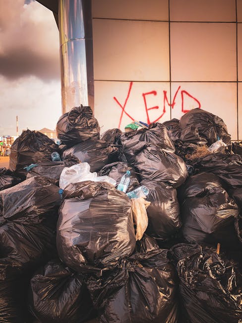 A large pile of black plastic rubbish bags, some appearing slightly crumpled, stacked against a modern tiled building wall. A few discarded plastic bottles and white waste materials are visible among the bags. The bags occupy the foreground and partially obscure the lower part of the wall, which features large beige tiles with thin grout lines. In the background, part of a cloudy sky is visible with a hint of distant cityscape, including a pole or antenna. The scene suggests an instance of private waste accumulation, possibly awaiting collection or disposal, which aligns with independent rubbish removal or on-site clearance services provided by companies like House Clearance Bromley. The overall atmosphere is ordinary and neutral, with natural lighting emphasizing the textures of the waste bags and the building’s reflective surface. The graffiti on the tiled wall contains the red spray-painted word 'XEND', adding an urban element to the setting and subtly highlighting the context of non-authority waste handling.