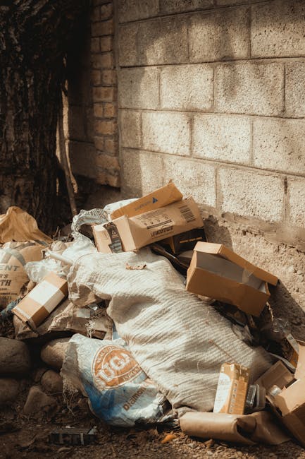 On a paved sidewalk adjacent to a black metal fence and a green leafy hedge, there are three large, tightly filled waste bags made of black plastic and white plastic material. The bags appear to contain household rubbish or garden waste, with some text or branding visible on the white bag. They are positioned close to the curb, with two standing upright and one lying against them, all slightly leaning to one side. The environment appears to be an outdoor residential area, with faint outlines of trees and a building or structure visible beyond the fence. The scene is captured in natural daylight with soft shadows, indicating an overcast or partly cloudy sky. This setting aligns with private rubbish collection or on-site clearance activities often handled by waste management services such as House Clearance Bromley, serving local house clearance or rubbish removal needs in Bromley or surrounding areas.