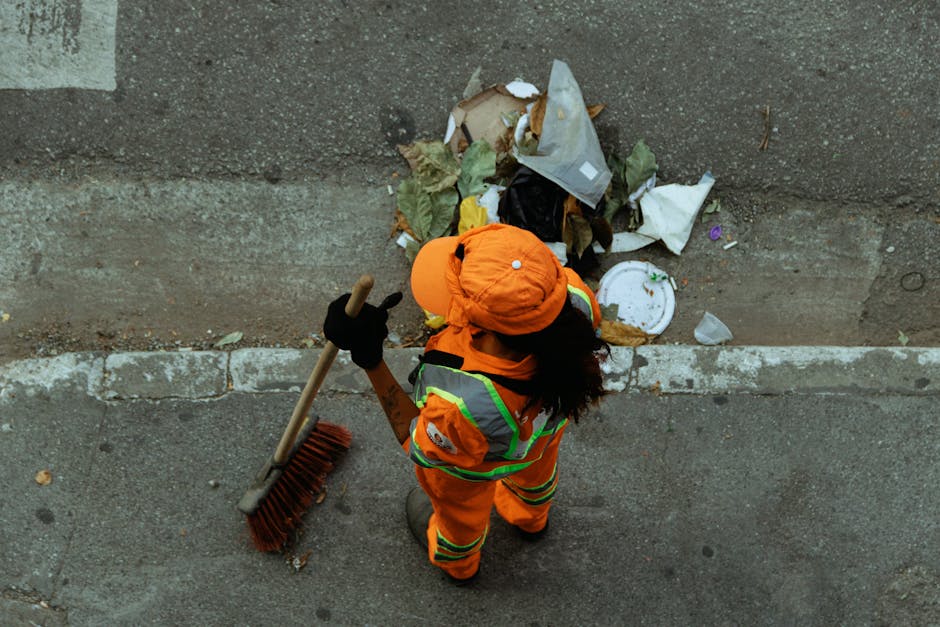 A worker dressed in high-visibility orange clothing with reflective yellow and silver strips, wearing a matching orange hard hat and black gloves, is standing on a paved sidewalk next to a low concrete wall. The worker is holding a broom with a wooden handle and red bristles. In front of them, there is a pile of mixed rubbish, including crumpled paper, plastic bottles, a food container, and other debris, situated on the ground and partially against the wall. The scene is outdoors, with diffuse natural lighting, and appears to capture a routine street cleaning activity that may be related to private waste management services such as those offered by House Clearance Bromley, aligning with themes of rubbish removal and on-site clearance in urban residential areas.