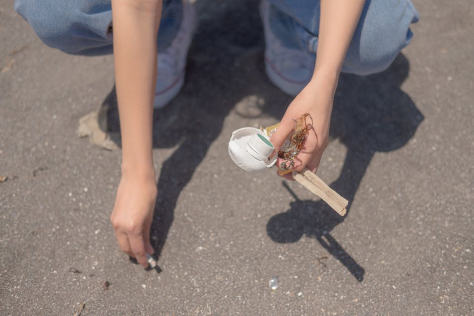 A person wearing light blue jeans and white sneakers is crouched down on a paved surface, holding a small collection of household waste items in their right hand, including a broken white ceramic or plastic object, some tangled paper or string, and a wooden stick or piece of scrap wood. Their left hand is resting on the ground beside them. The background shows the asphalt surface with small scattered debris, indicating an outdoor setting likely in a driveway or parking area. The image emphasizes the visual task of collecting and disposing of small waste items, relevant to independent rubbish removal or site clearance services provided by companies like House Clearance Bromley. The lighting is natural, and the scene captures a moment of manual waste tidying, supporting the theme of alternative waste handling outside of conventional waste collection systems.