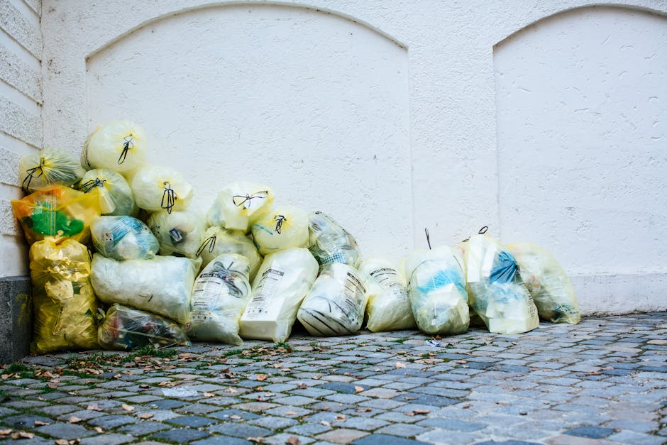 A pile of discarded cardboard boxes, some flattened and others still in their packaging, stacked unevenly against a weathered brick wall with visible mortar joints. The boxes vary in size, with printed labels and barcodes visible on some, and are mixed with crumpled brown paper and plastic packaging materials. A large, textured fabric sack is partially buried among the debris, showing signs of wear and dirt, suggesting it has been used for waste collection. The surrounding ground consists of dirt and small rocks, with a portion of a tree trunk visible on the left side of the image. The background features a slightly sunlit outdoor setting near a building or boundary wall, indicative of an area where waste is temporarily stored before removal, aligning with the concept of private or alternative rubbish disposal services like those offered by House Clearance Bromley. The scene emphasizes the informal nature of debris accumulation that can result from last-minute rubbish clearance or home decluttering efforts, requiring professional waste removal solutions.